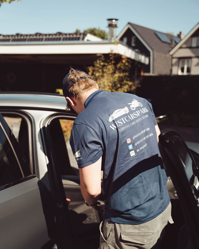 Ruben aan het werk bij het open portier van een grijze BMW, met het WestCarSpark-logo op zijn rug