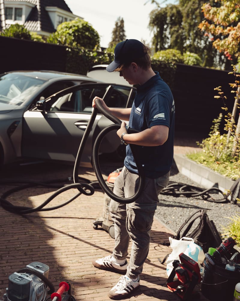 Ruben in een WestCarSpark-poloshirt rolt de stofzuigerslang op naast een grijze BMW